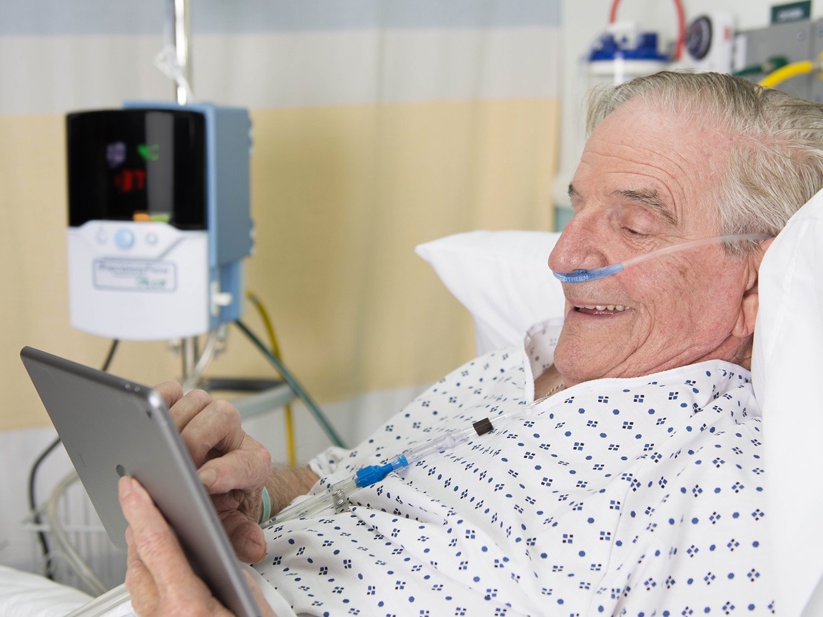 Image of elderly male patient with a Vapotherm cannula smiling at a tablet he’s using. A Vapotherm Precision Flow is visible in the background.
