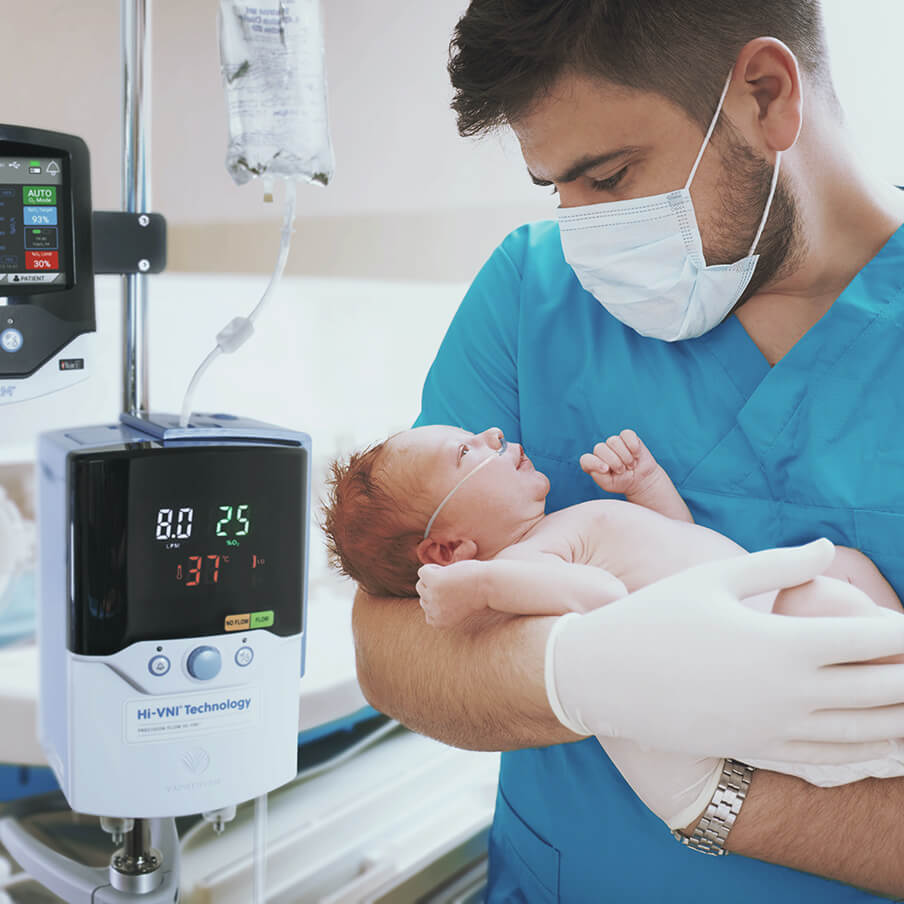 A masked male clinician in scrubs and gloves holding a neonate